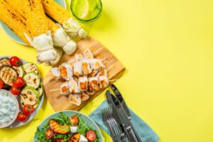 Bird's eye view of corn on the cob, grilled chicken, vegetables, and silverware on a yellow surface